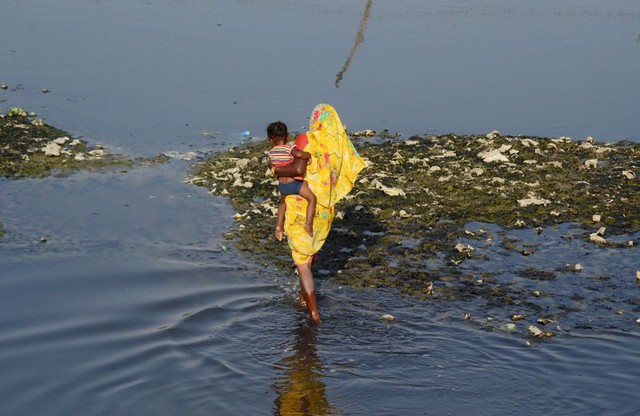  Một người phụ nữ Ấn Độ bồng con, đi chân trần trên một đoạn sông ô nhiễm 