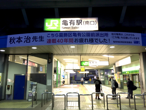 
Một tấm biểu ngữ trắng lớn được đặt ở ngay trước cửa ra vào nhà ga Kameari với lời cảm ơn và chúc mừng tác giả Akimoto Osamu vì 40 năm làm việc cống hiến hoàn thành bộ manga
